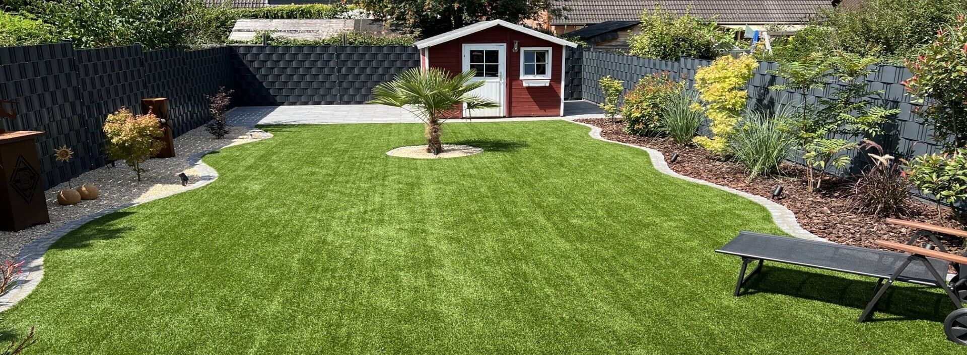 Von Kalla Gartenbau in Willich gestalteter Garten mit Rasenfläche, Palme und rotem Gartenhaus